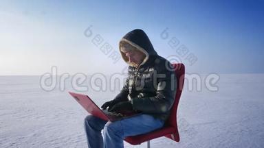穿着温暖外套和引擎盖的商人坐在雪地沙漠里，用笔记本电脑工作，转向相机，兴高采烈地微笑着。